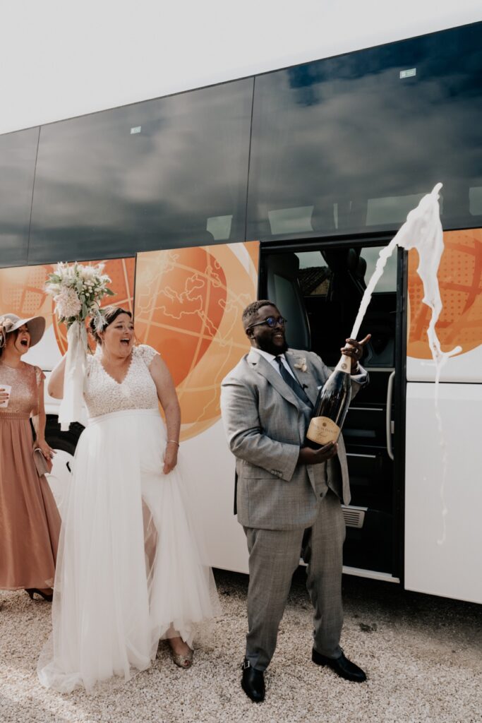 un couple ouvre une bouteille de champagne devant le bus qui a ramené leurs invités. Capturé par les photographes de mariage Céline et Thibaut Deligey