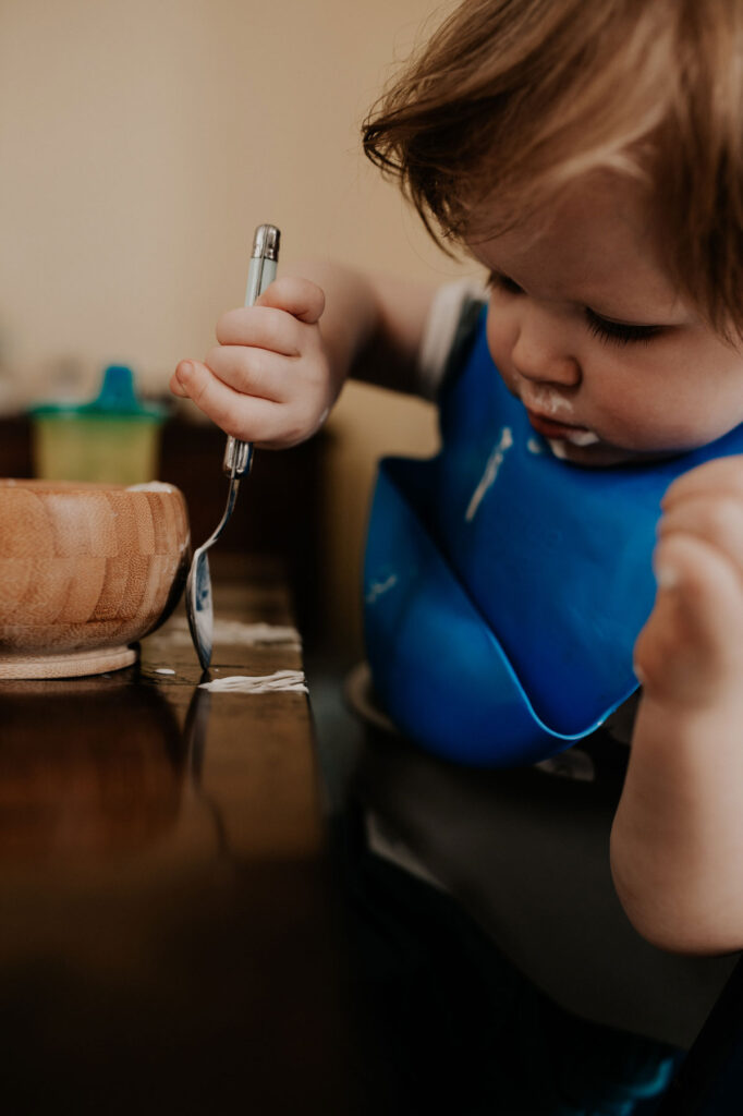 un petit garçon mange tout seul comme un grand. il essaye de rattraper du yaourt sur la table avec sa cuillere.