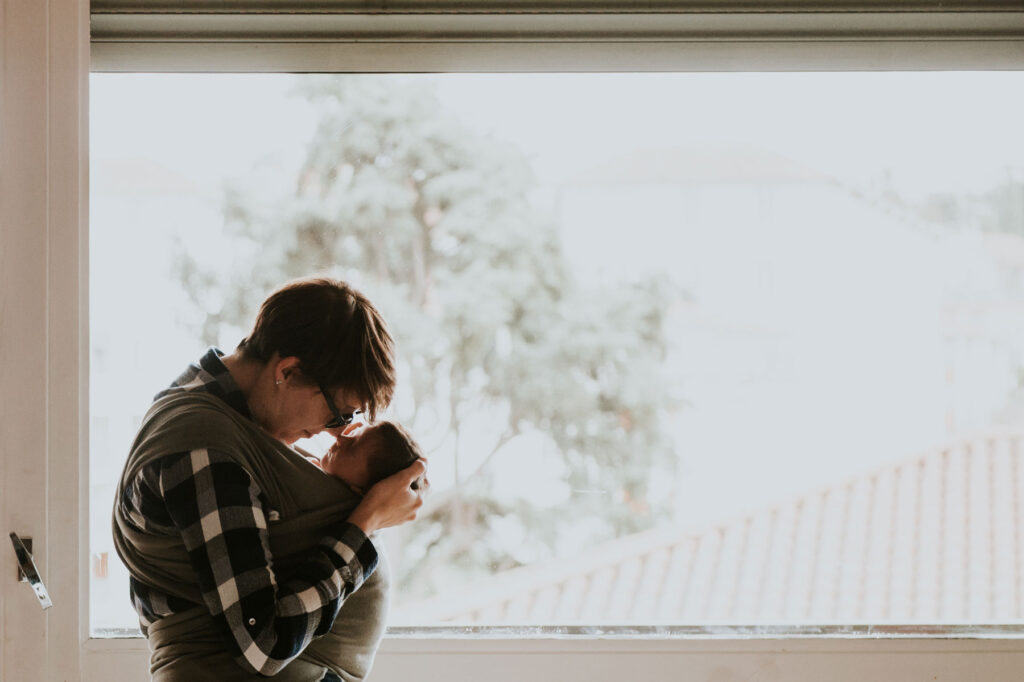 calin maman et bébé en porte bébé devant une fenetre