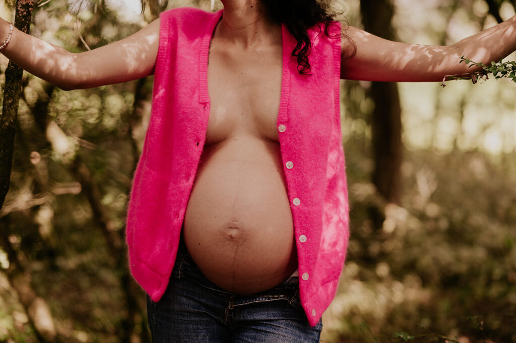 séance photo de grossesse. une femme enceinte porte un gilet rose, en exterieur, ventre nu.