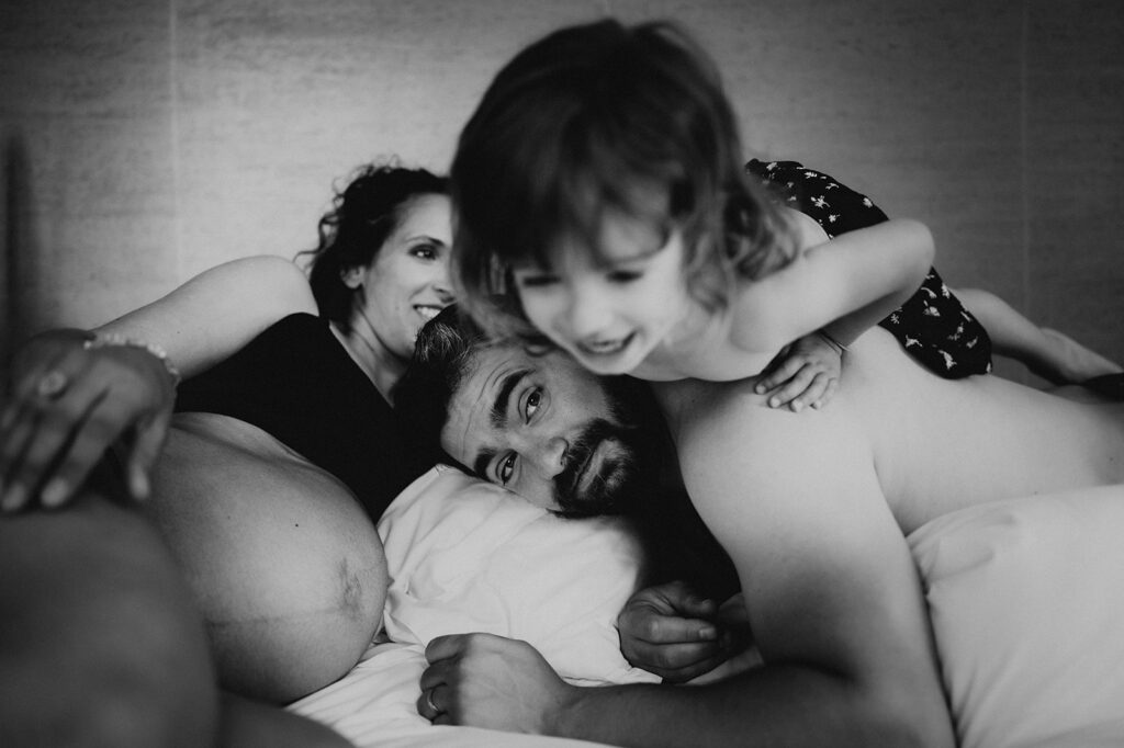 photo en noir et blanc issu d'une séance grossesse. la femme enceinte le papa et la grande soeur dans un lit