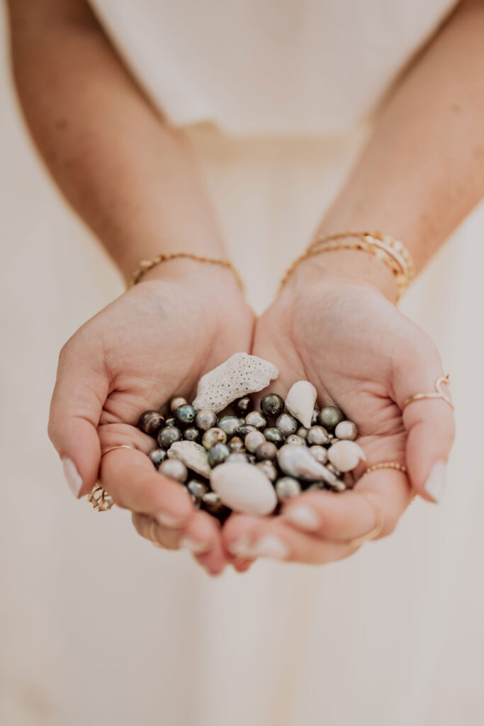 Mains féminines tenant des perles noires de Tahiti et des morceaux de corail blanc, photographie poétique et délicate en Polynésie française.